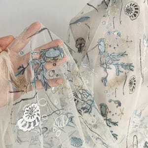 Può includere: Tessuto bianco trasparente con ricami di motivi marini. Il ricamo presenta conchiglie, stelle marine, cavallucci marini e alghe in tonalità blu e oro. Il tessuto è sollevato, mostrando i delicati dettagli del design.