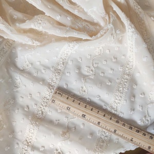 May include: Ivory white sheer fabric with a textured, raised dot pattern and embroidered white floral designs. A wooden ruler with centimeter markings is placed on the fabric.