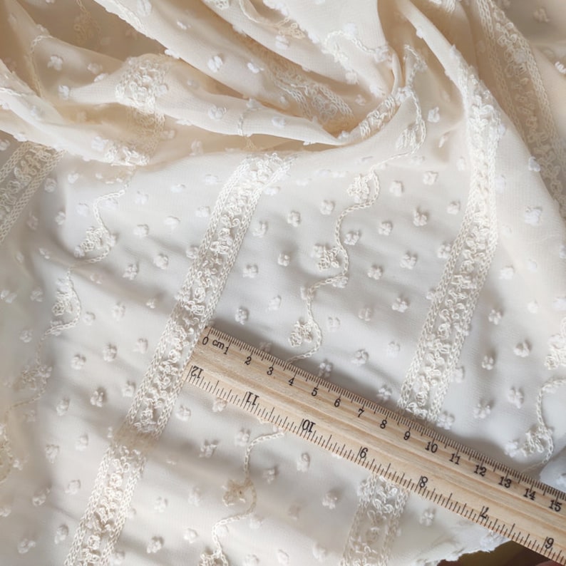 May include: Ivory white sheer fabric with a textured, raised dot pattern and embroidered white floral designs. A wooden ruler with centimeter markings is placed on the fabric.