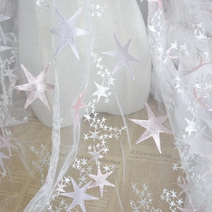May include: White tulle fabric with pink and white embroidered stars. The fabric is sheer and delicate, perfect for crafting or decorating.