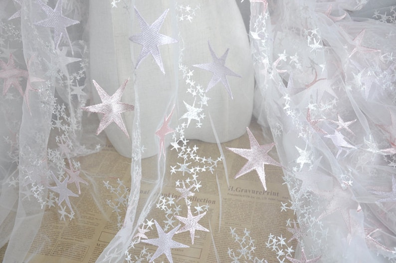 May include: White tulle fabric with pink and white embroidered stars. The fabric is sheer and delicate, perfect for crafting or decorating.