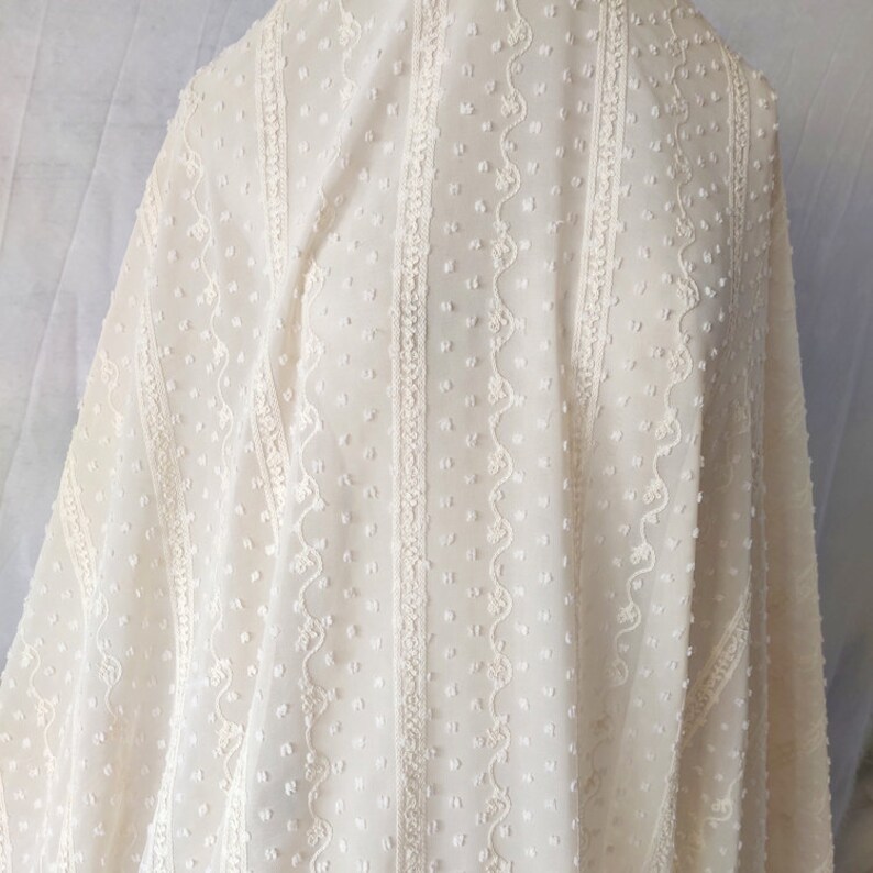 May include: White sheer fabric with a dotted and embroidered pattern. The fabric has vertical stripes of embroidery with a scalloped design.