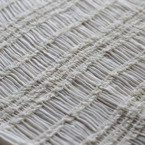 May include: White fabric with a textured, gathered, or smocked pattern. The fabric has a slightly shiny finish.