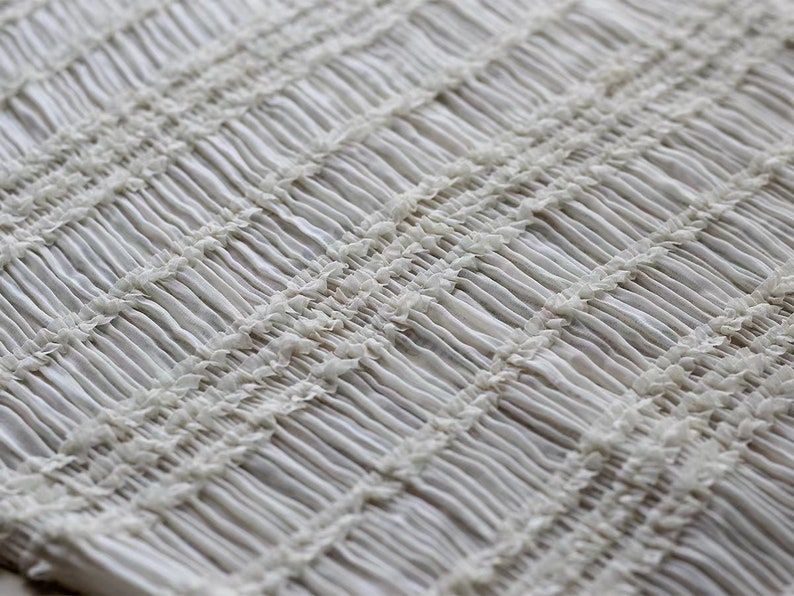 May include: White fabric with a textured, gathered, or smocked pattern. The fabric has a slightly shiny finish.