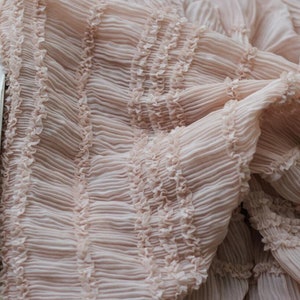 May include: A close-up of a pale pink fabric with a ruffled texture. The fabric is folded and draped, showing the intricate details of the ruffles.