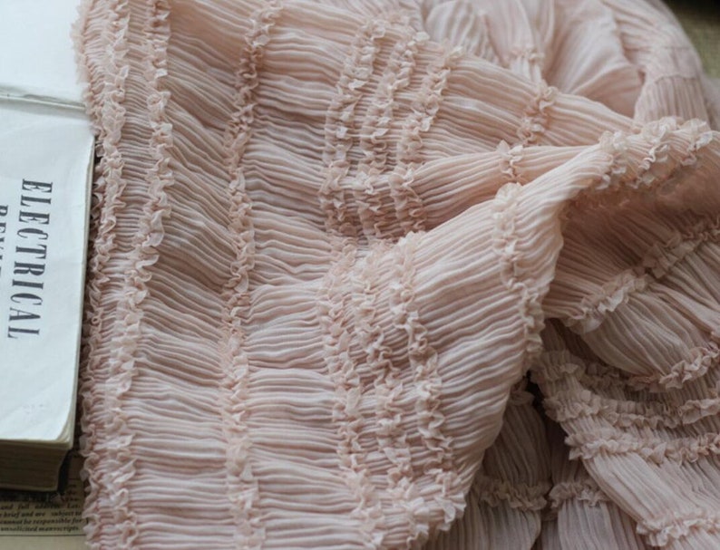 May include: A close-up of a pale pink fabric with a ruffled texture. The fabric is folded and draped, showing the intricate details of the ruffles.
