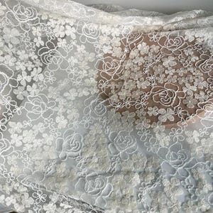 May include: White floral embroidered tulle fabric with a delicate pattern of small flowers and larger roses. The fabric is sheer and lightweight, making it ideal for crafting, sewing, and decorating.