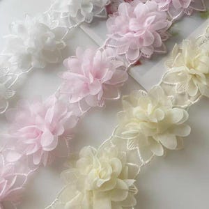 Könnte beinhalten: Nahaufnahme von floraler Spitzenborte in Weiß, Hellrosa und Blassgelb. Die zarten Blüten sind mit transparenten Blütenblättern geschichtet und an einer Borte mit Muschelkanten befestigt. Diese Borte eignet sich für verschiedene Bastel- und Nähprojekte.