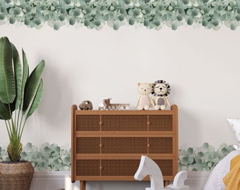 May include: A white wooden rocking horse with a brown wooden chest of drawers with three drawers. The chest of drawers has a woven rattan front. The wall behind the furniture is white with a green eucalyptus leaf garland along the top and bottom of the wall. There is a green potted plant in the corner of the room.