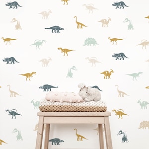May include: A white wall with a repeating pattern of dinosaur silhouettes in various colours, including blue, green, brown, and beige. A small wooden stool with a knitted throw and a stuffed dinosaur toy sits in front of the wall.