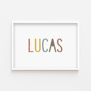 May include: A white framed print with the name "LUCAS" written in colorful, hand-drawn letters. The letters are red, yellow, blue, brown, and red.