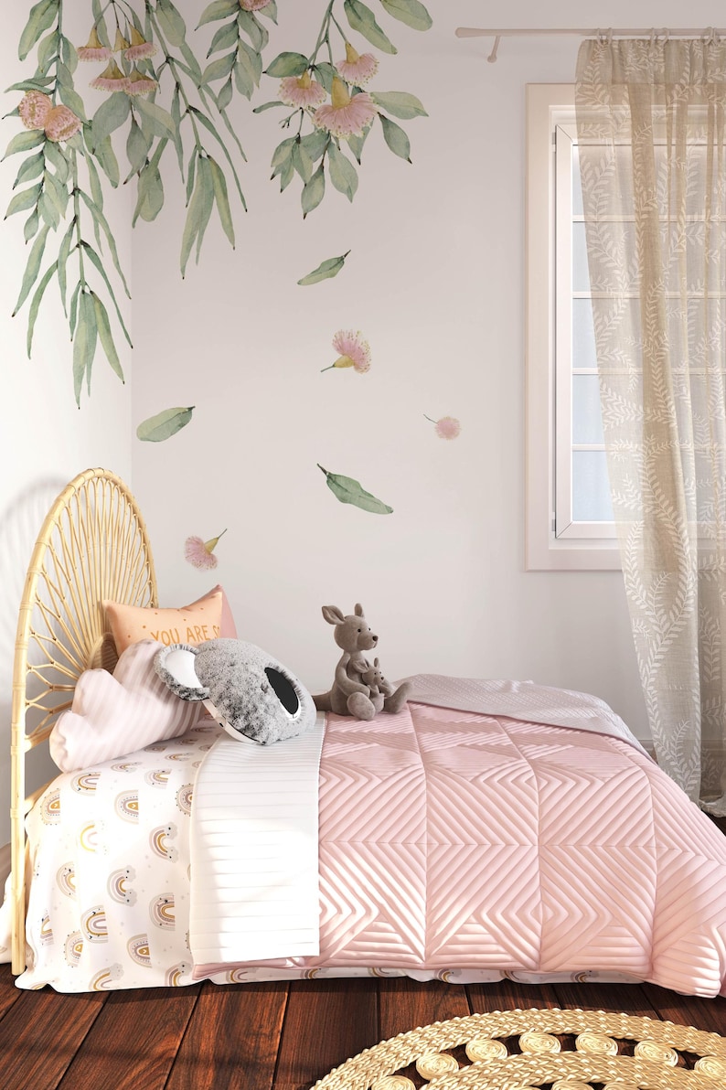 May include: A pink and white bed with a white and gold rainbow patterned sheet and a pink quilted blanket. The bed has a wicker headboard and a stuffed kangaroo and koala on the bed. The wall behind the bed has a floral decal with pink flowers and green leaves.