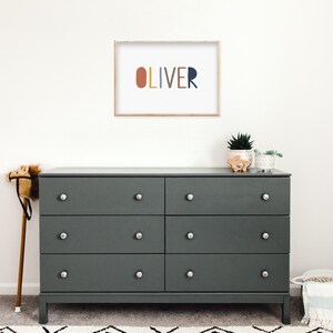 May include: A dark gray dresser with six drawers and silver knobs. The dresser is topped with a framed print that says "OLIVER" in colorful letters. There are also two small potted plants and a wooden toy on top of the dresser.