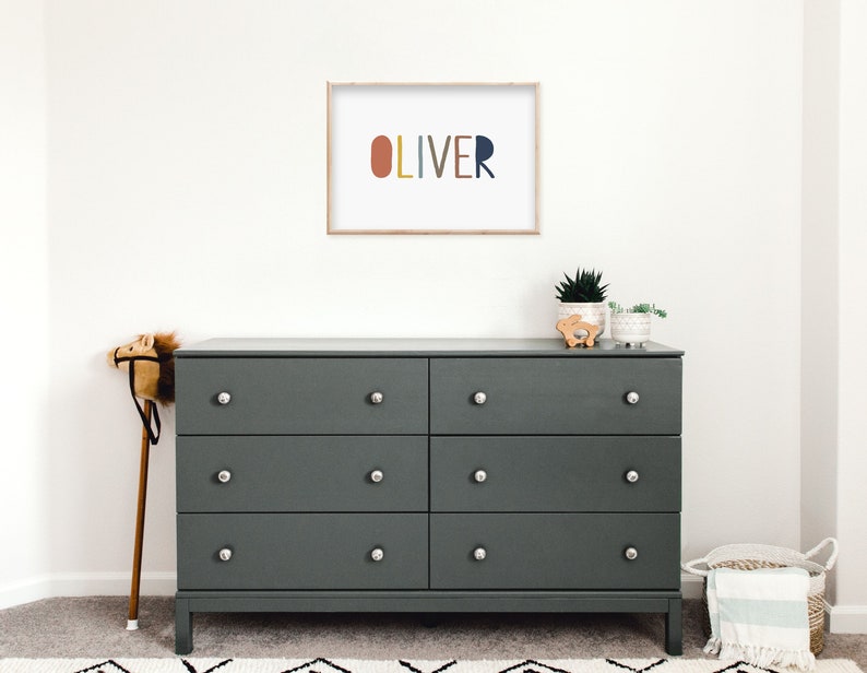 May include: A dark gray dresser with six drawers and silver knobs. The dresser is topped with a framed print that says "OLIVER" in colorful letters. There are also two small potted plants and a wooden toy on top of the dresser.