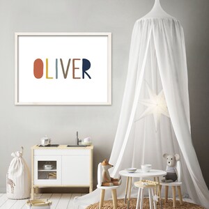May include: A white framed print with the name "OLIVER" spelled out in colorful, rounded letters. The print is hanging on a light gray wall in a room with a white canopy, a play kitchen, and a small table and chairs.