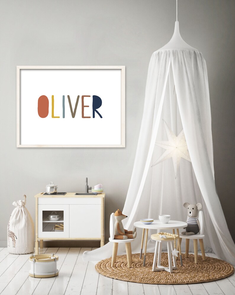 May include: A white framed print with the name "OLIVER" spelled out in colorful, rounded letters. The print is hanging on a light gray wall in a room with a white canopy, a play kitchen, and a small table and chairs.