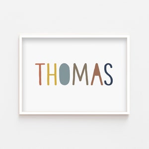 May include: A white framed print with the name "THOMAS" written in colorful, stylized letters. The letters are in shades of red, yellow, blue, brown, and gray.