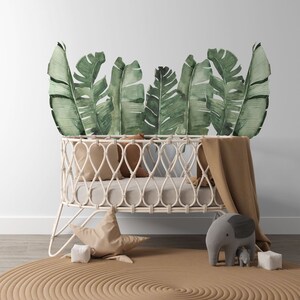 May include: A white wicker cot with a brown blanket and a plush grey elephant toy. The cot is decorated with a wall decal of green banana leaves.