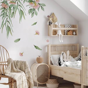 May include: A white wall with a floral decal featuring pink flowers and green leaves. A wooden bed with a white and black patterned pillow is in the foreground. A wicker side table with a woven basket is next to the bed.
