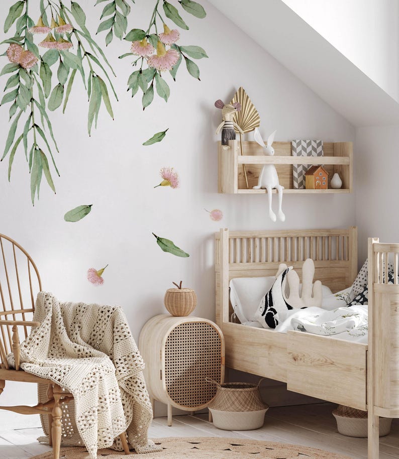 May include: A white wall with a floral decal featuring pink flowers and green leaves. A wooden bed with a white and black patterned pillow is in the foreground. A wicker side table with a woven basket is next to the bed.