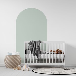 May include: A white cot with a grey knitted blanket and a brown throw blanket. The cot is in front of a white wall with a light green arch painted on it. There is a wicker basket and a world map rug on the floor.