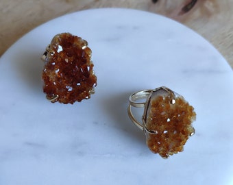 May include: Two gold rings with orange citrine crystal clusters. The rings are on a white marble surface.