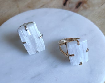 May include: Two selenite crystal rings, one with a silver band and one with a gold band. The rings are on a white marble surface.