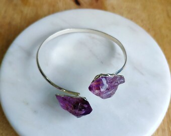 May include: A silver bangle bracelet with two amethyst crystal accents. The crystals are a deep purple color and have a natural, rough texture.
