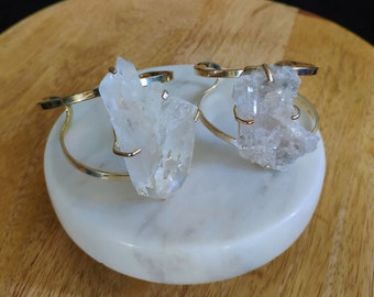 May include: Two gold wire bracelets with clear quartz crystals attached. The bracelets are on a white marble surface.