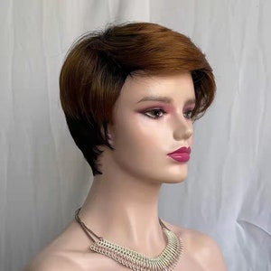 May include: A mannequin head displaying a short, layered wig with a brown and black ombre effect. The mannequin also wears a silver-toned necklace with a unique, layered design. The background is a plain white sheet.