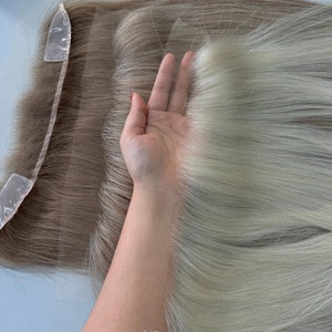 May include: A light blonde human hair topper with a lace front. The topper is laid flat on a table and a hand is holding the topper up to show the length of the hair. The text "INE HAIRLINE TOPPER" is printed on the table.