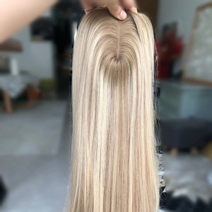 May include: A long, blonde hairpiece with a natural-looking parting. The hair is a light, golden blonde colour and appears to be straight. The hairpiece is held up by a hand, showcasing the top and the flowing strands.