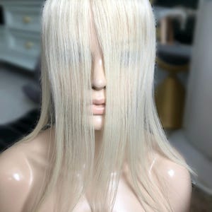 May include: A mannequin head with a pale blonde wig. The wig features long, straight hair that covers the face. The mannequin has a neutral expression and is set against a blurred background.
