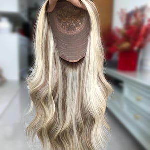 May include: A blonde wig with long, wavy hair. The wig features highlights of light brown and blonde. The cap is a light brown color with a lace design. The wig is held up by a hand.