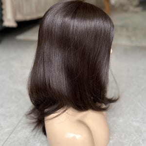 Op de afbeelding: Een donkerbruine pruik met een stompe snit, gepresenteerd op een mannequinhoofd. Het haar is steil en glanzend, met licht gelaagde uiteinden. De pruik is ontworpen om te worden gedragen en gestyled.