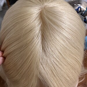 Puede incluir: Vista cercana de la parte superior de una peluca rubia. La peluca est&aacute; hecha de cabello sint&eacute;tico y tiene una raya de aspecto natural.