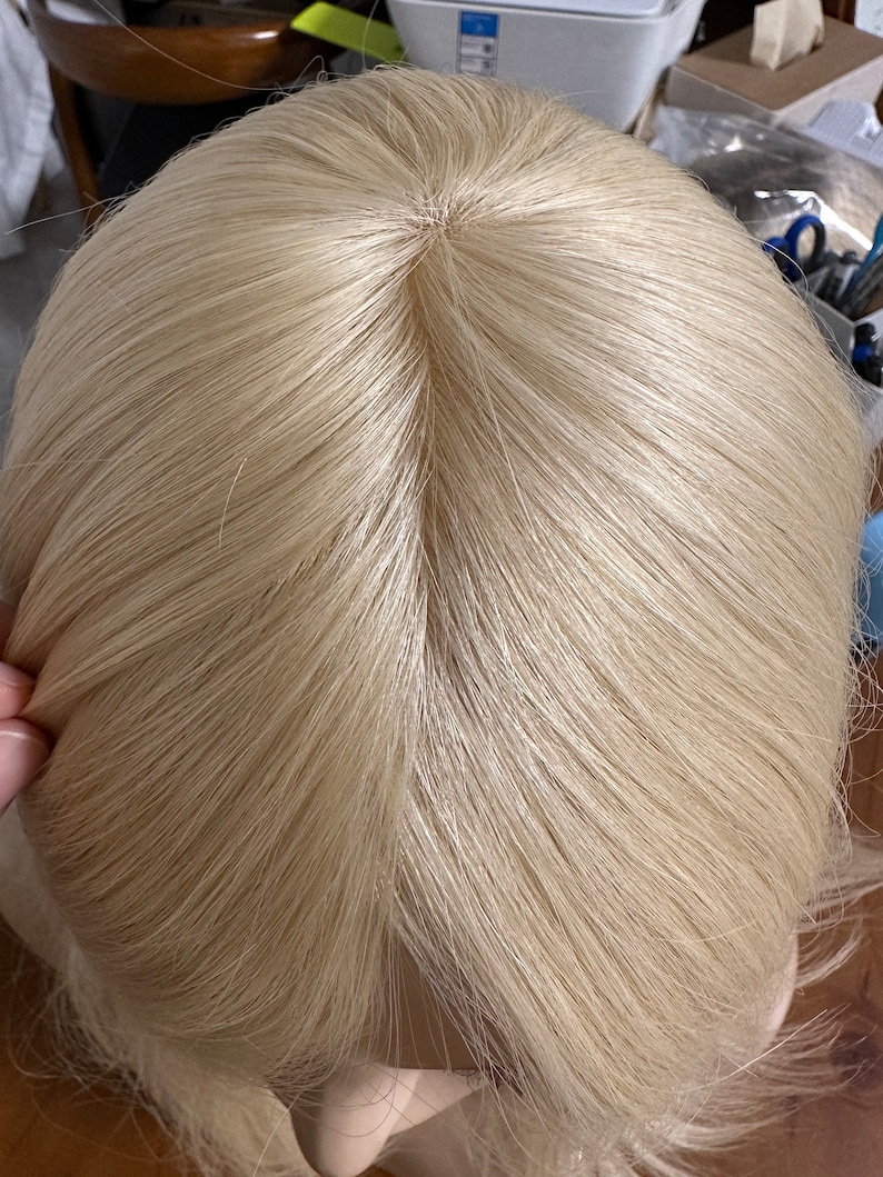 Puede incluir: Vista cercana de la parte superior de una peluca rubia. La peluca est&aacute; hecha de cabello sint&eacute;tico y tiene una raya de aspecto natural.