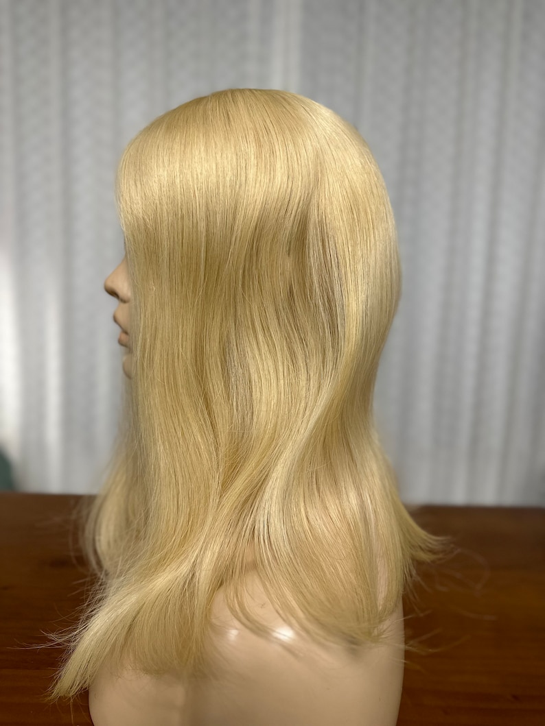 Puede incluir: Una peluca larga, lisa y rubia en una cabeza de maniqu&iacute;. La peluca est&aacute; peinada con raya lateral y capas.