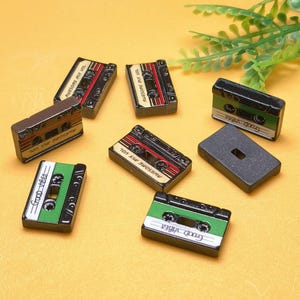 May include: A collection of miniature cassette tape replicas in various colors. Some tapes feature the text "AWESOME MIX VOL" and "GOOD VIBES". The tapes have a retro aesthetic, with red, green, and black accents, and are arranged on a yellow surface.