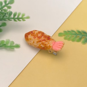 May include: A novelty hair clip shaped like a fried shrimp. The clip features a light brown body with reddish-brown details and a pink tail. The clip is attached to a small silver-colored metal clasp.