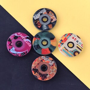 Puede incluir: Un conjunto de cinco rodamientos de ruedas de monopatín coloridos. Cada rodamiento presenta un diseño único con texto y patrones en varios colores, incluyendo rosa, verde, naranja y azul. El texto incluye frases como "This is life" y "Fashion".