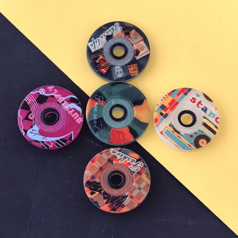 May include: A collection of five colorful skateboard wheel bearings. Each bearing features a unique design with text and patterns in various colors, including pink, green, orange, and blue. The text includes phrases like "This is life" and "Fashion".
