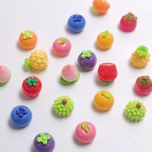 May include: A collection of miniature, colorful fruit-shaped charms. The assortment includes blueberries, strawberries, peaches, oranges, and pineapples. Each charm is brightly colored and detailed, with green accents. The charms are scattered on a white surface.