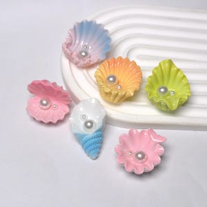 May include: A selection of colourful, seashell-shaped decorative items. The shells are in various colours, including pink, yellow, blue, and green, each with a pearl accent. The items are designed to resemble seashells.