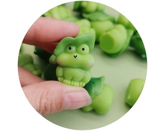 Puede incluir: Una pequeña figura de rana verde con un sombrero de hoja. La rana tiene ojos negros y una pequeña sonrisa. La figura está hecha de un material suave y blando.