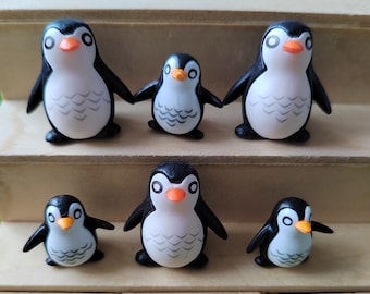 May include: Six black and white penguin figurines with orange beaks and black eyes. The penguins are arranged in two rows on a wooden surface.