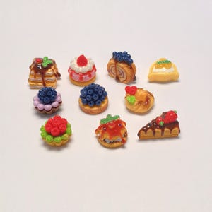 Puede incluir: Una colección de pasteles en miniatura y coloridos, que incluyen panqueques con jarabe, pasteles cubiertos de bayas y una rebanada de gofre cubierto de chocolate con una fresa. Los postres están dispuestos sobre una superficie blanca.