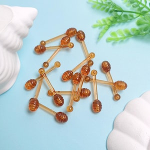 Könnte beinhalten: Eine Ansammlung kleiner, bernsteinfarbener Honigdipper. Jeder Dipper hat einen gerippten, oval geformten Kopf und einen schlanken Griff mit einer kleinen Kugel am Ende. Die Dipper sind auf einer hellblauen Oberfläche verstreut, mit einem weißen Dekorationselement und grünem Laub im Hintergrund.