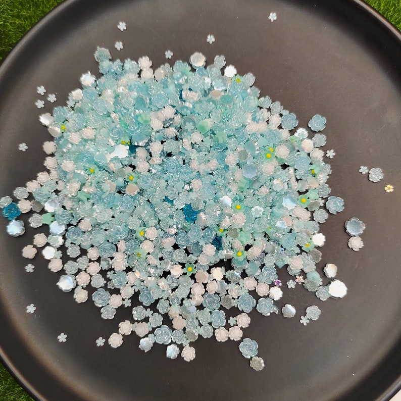May include: A close-up of a black plate filled with a variety of blue and white flower-shaped resin embellishments. The embellishments are scattered across the plate, creating a visually appealing and textured surface.