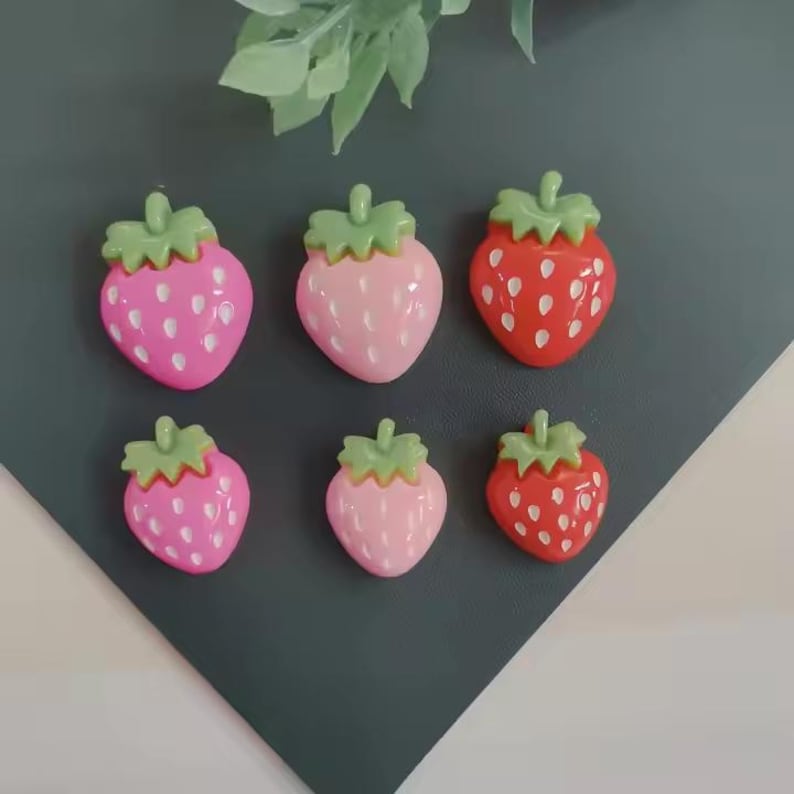 May include: Six strawberry-shaped decorative items. Two are bright pink, two are light pink, and two are red. Each has a green stem and white seed details. The items are arranged on a dark gray surface.